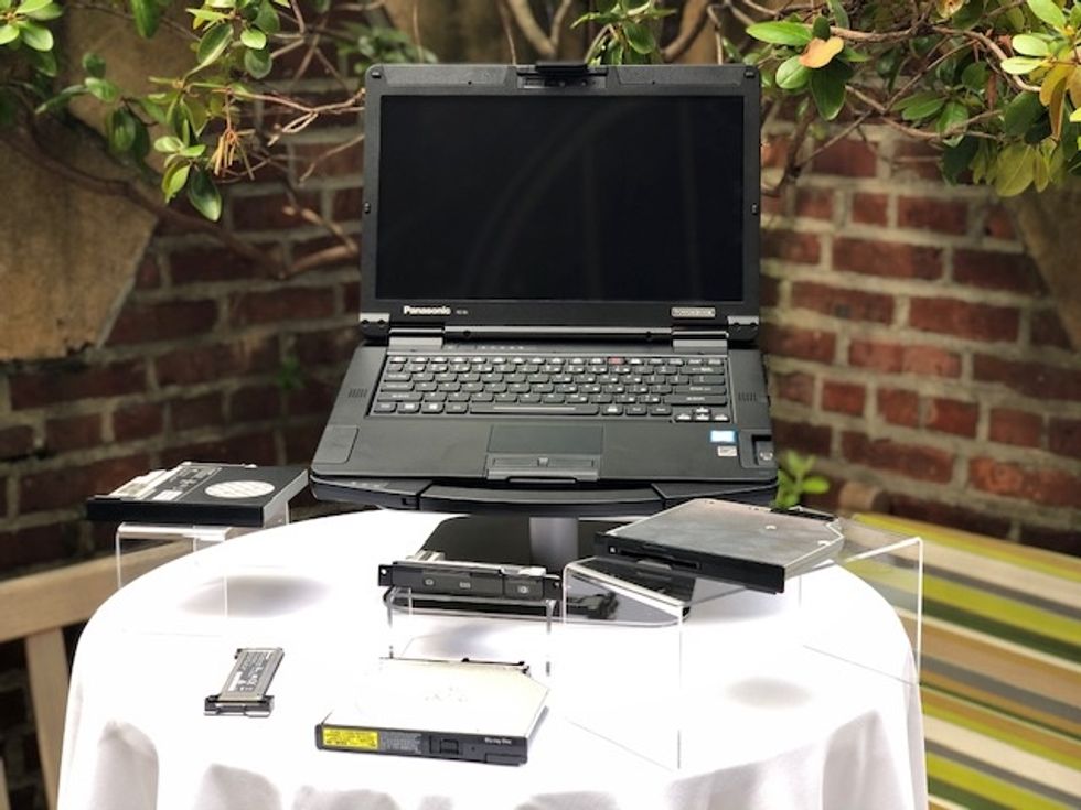 A black laptop with different drives around it, on a white table outside under a tree