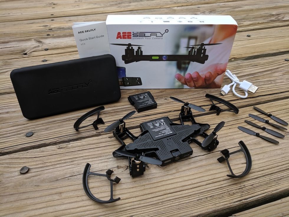 A drone, propellers, guards, cable, carrying box and a box that says 'AEE Selfly' seated on top of a wooden table