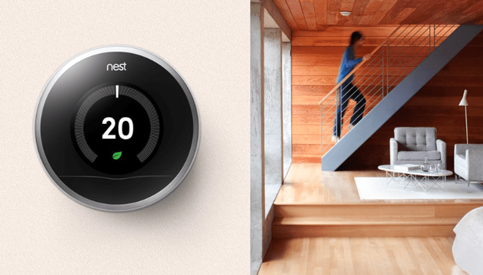 A Nest thermometer on a wall, reading 20 degrees Celsius, with a woman climbing a staircase at home in the background