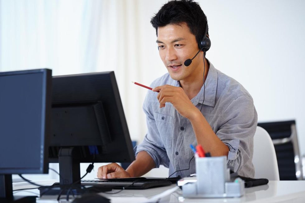 A photo of a man helping someone on a virtual call using his computer and a headset.