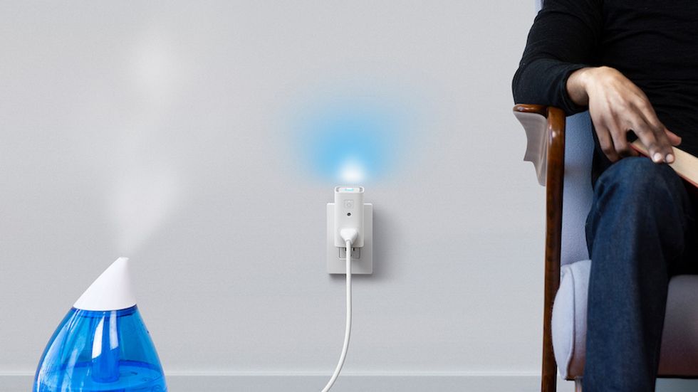 A rectangular plug in a wall outlet, with a blue light on top, and a small blue humidifier plugged with water vapor coming from the top next to a man sitting in a chair