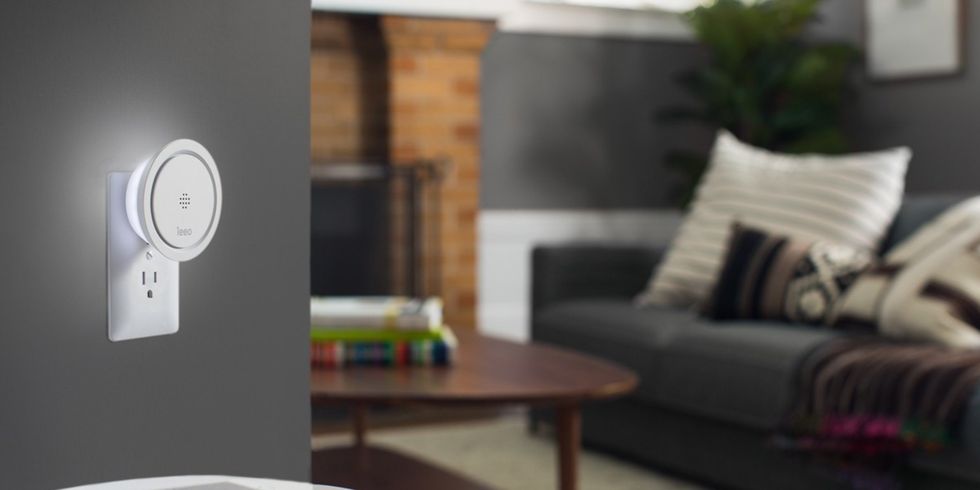 A white smart device from Leeo plugged into an outlet in a living room with a gray couch and white pillows