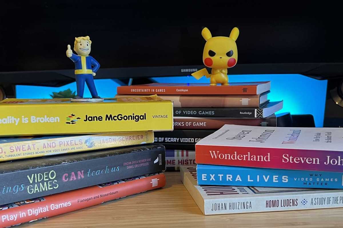 Gaming books stacked on a wooden desk with Fallout and Pokemon figures on top