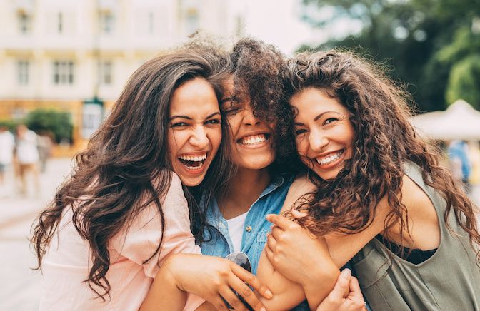 Three people with long brown curly hair smiling outside as their photo is being taken