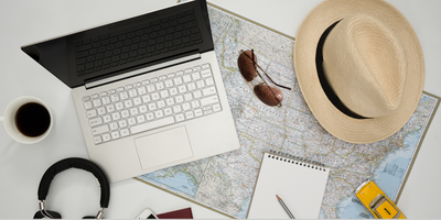 a photo of a laptop, cup of coffee, sunglasses, a hat, notebook and pen on top of a map on a desk.