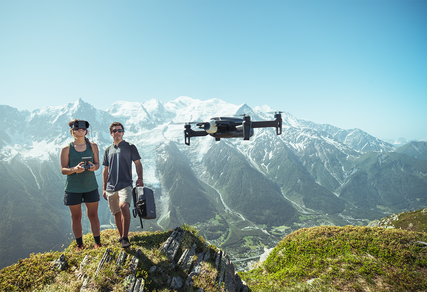 Two people flying a large drone in the foreground, with snow-capped mountains in the background