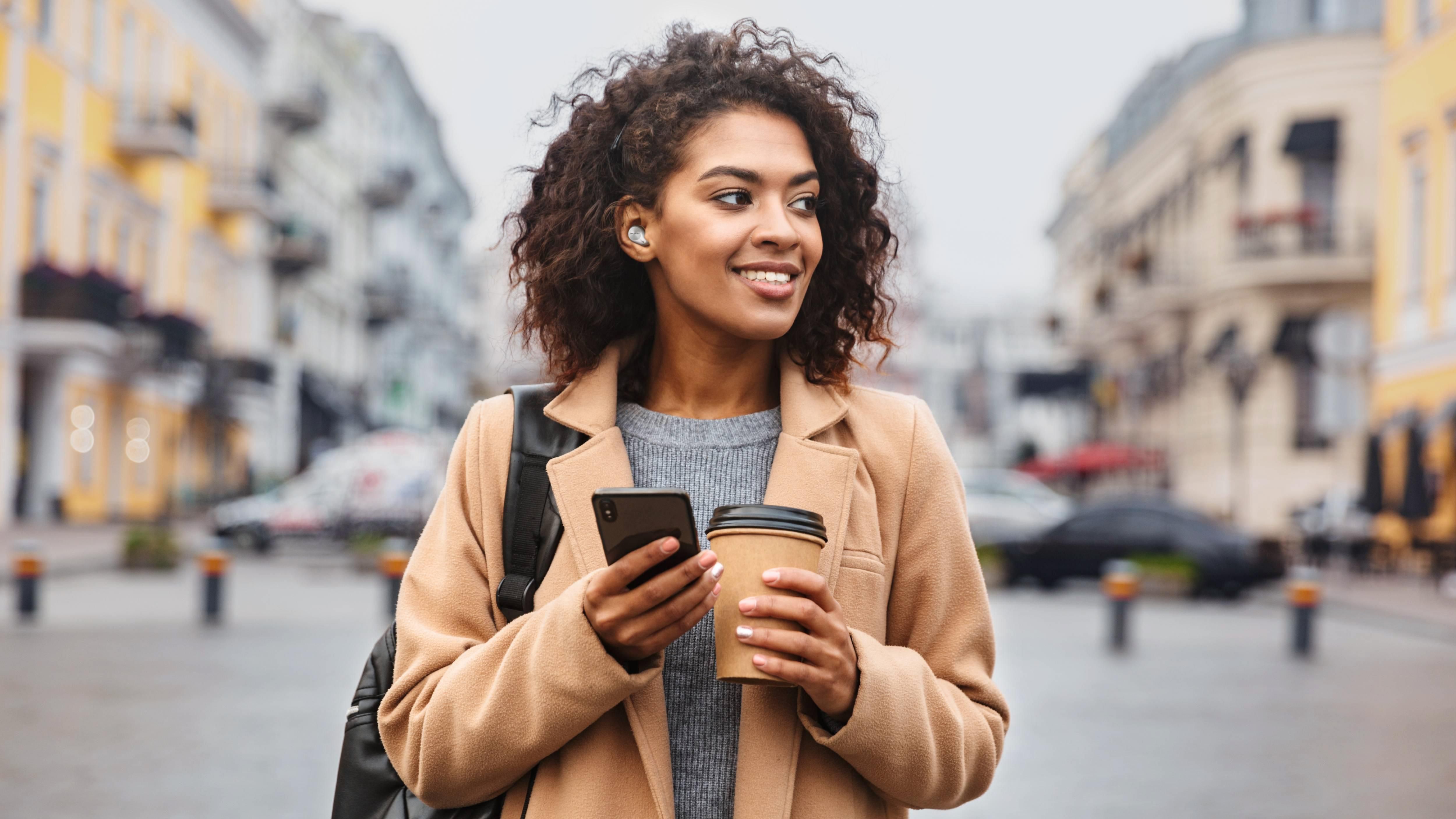 Woman wearing Technics earbuds and holding a coffee mug and smartphone