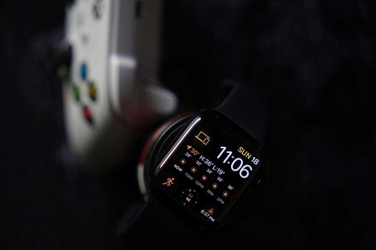 Close-up view of a smartwatch displaying time and weather next to a game controller on a dark background.