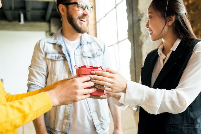 Three people exchanging gifts in an office space with a large window