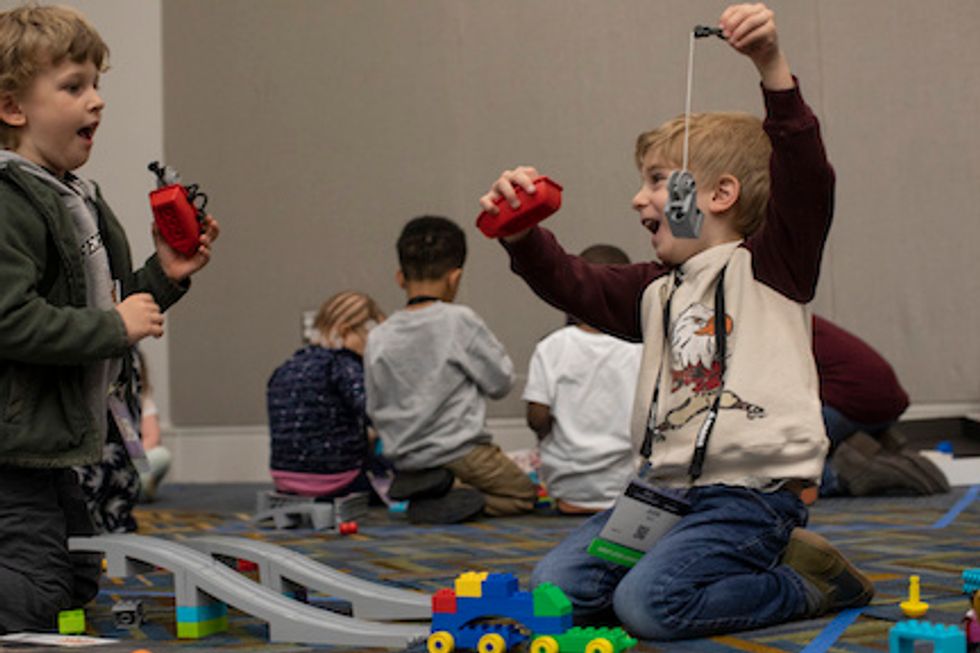Two young boys enthusiastically play with color plastic blocks, next to a plastic toy ramp, with other children playing nearby
